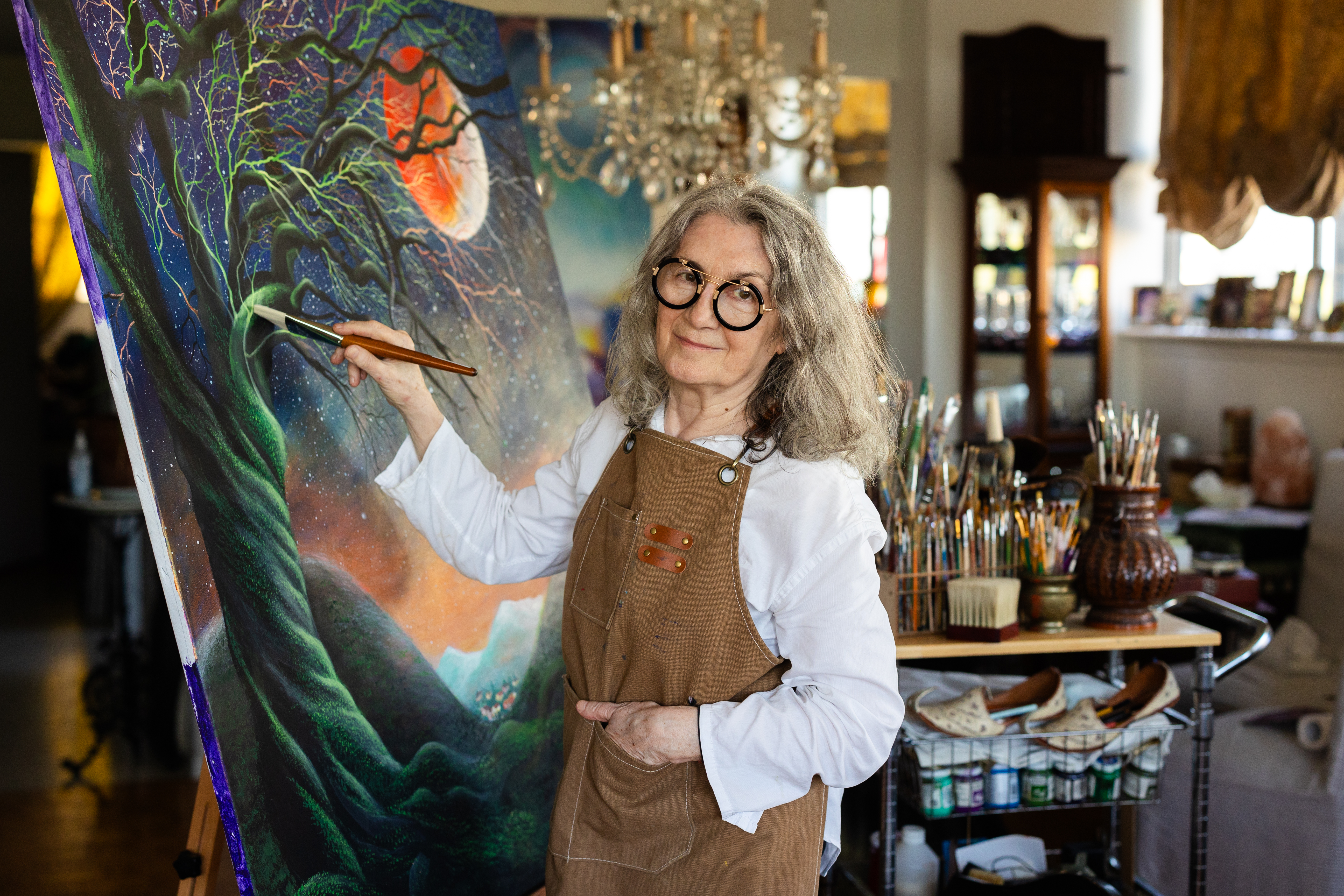 Tatjana Kilibarda seated in her studio beside a large painting, holding a paintbrush.